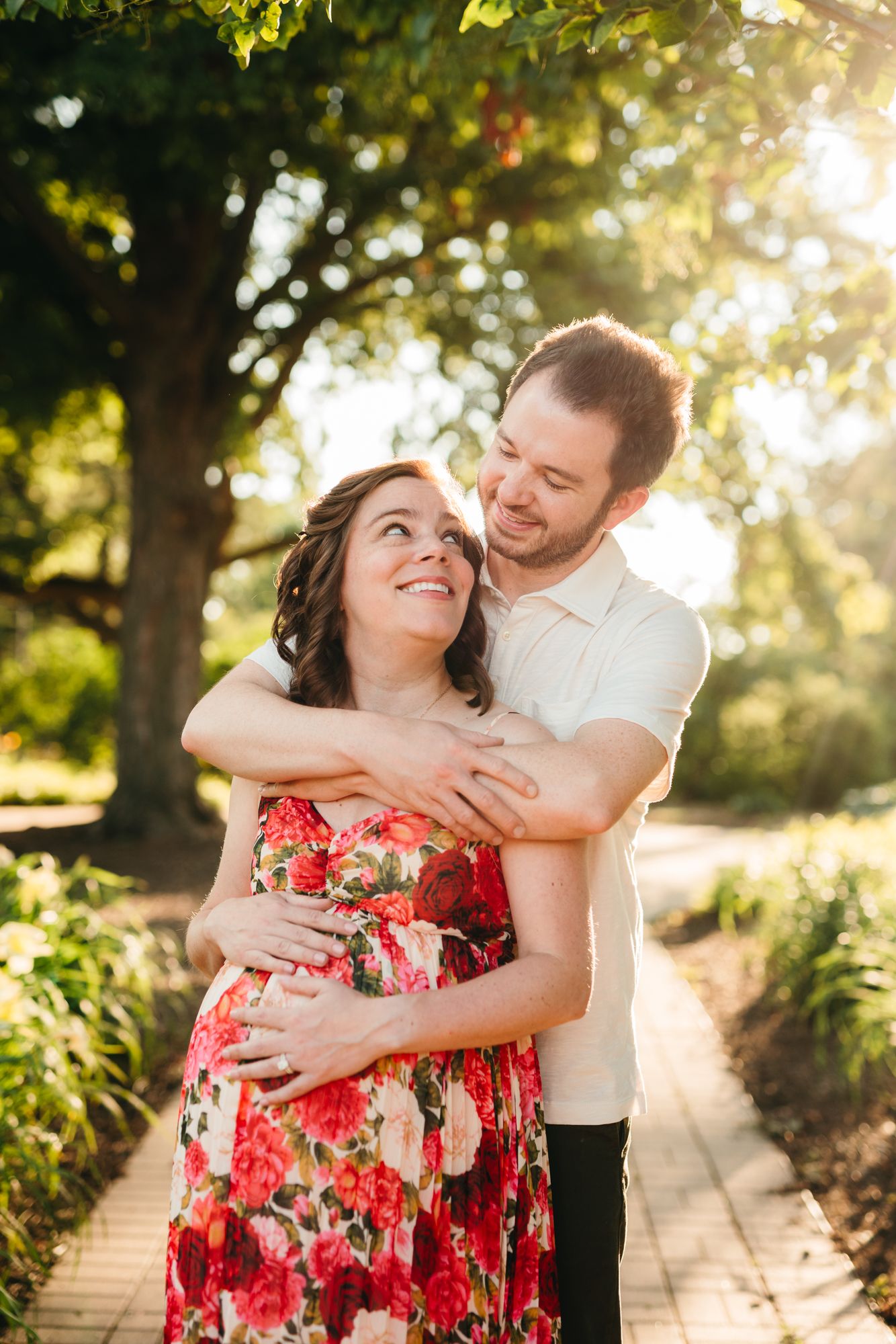 A smiling man embraces his pregnant partner in a floral dress from behind on a sunlit garden path lined with trees.