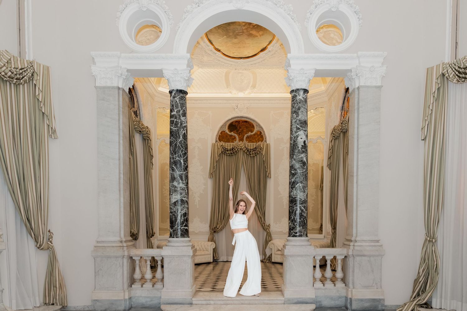 Bride in a white jumpsuit raises her arms in an ornate marble hall with arched columns, draped curtains and a frescoed ceiling.