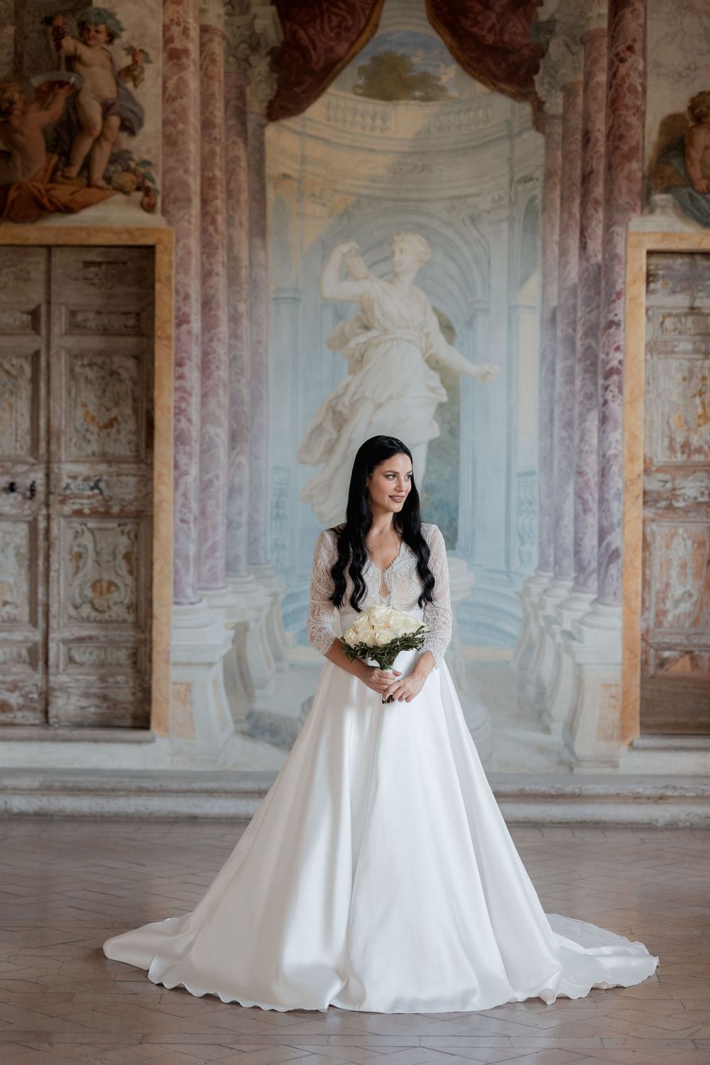 Bride in a white lace-sleeve satin ball gown holding a white bouquet in a frescoed Italian palace hall with painted statue and columns.