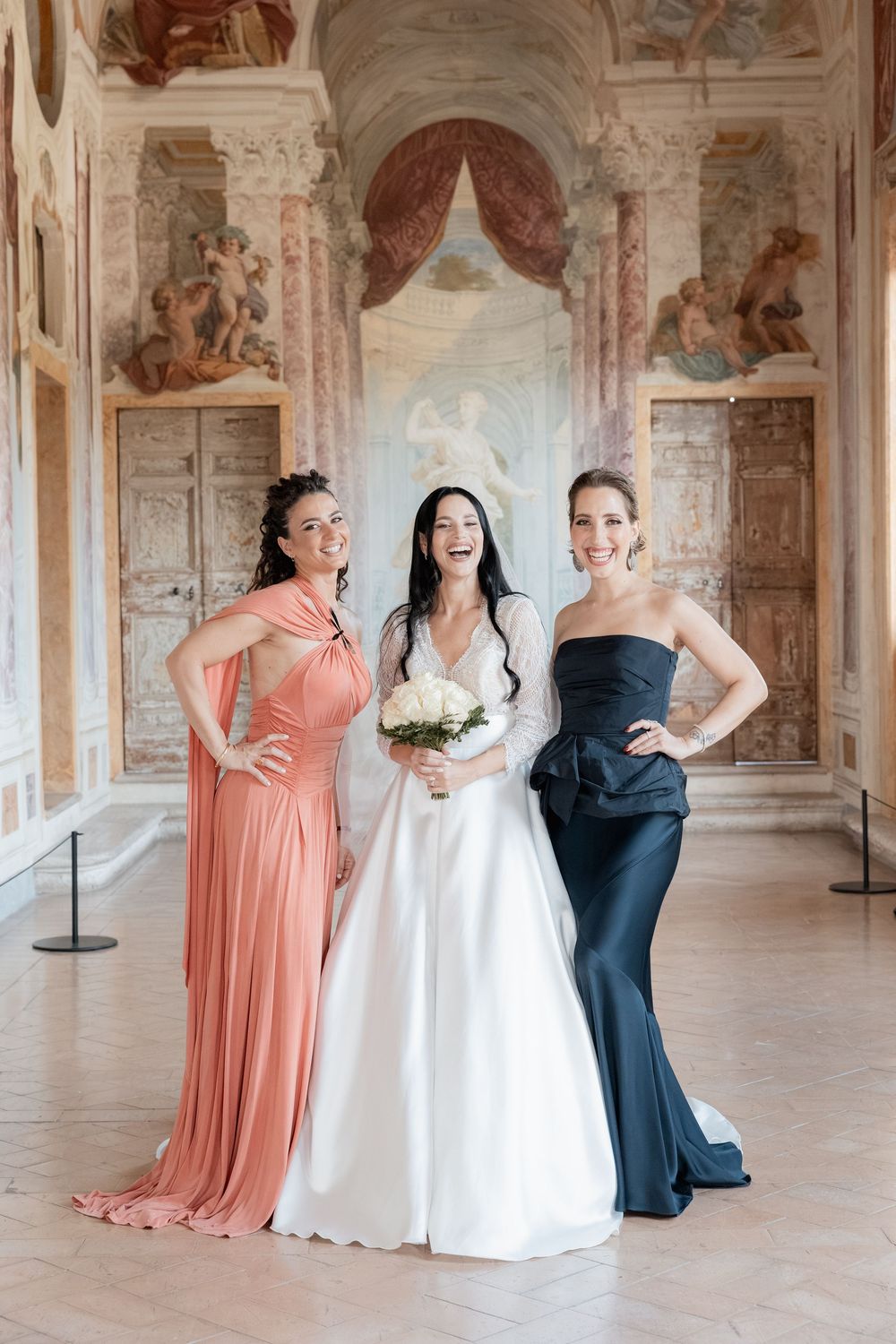 Smiling bride in a white gown holding a bouquet poses with two bridesmaids in coral and navy dresses inside a frescoed Italian palace hall.