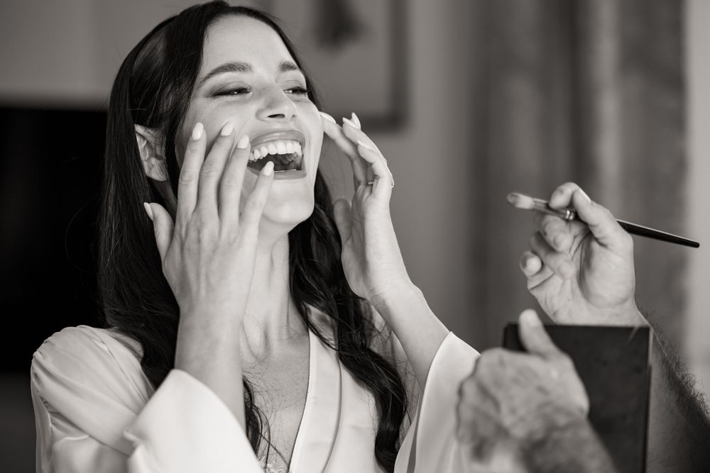 Black and white photo of a laughing woman in a robe with long dark hair as a makeup artist applies makeup with a brush.