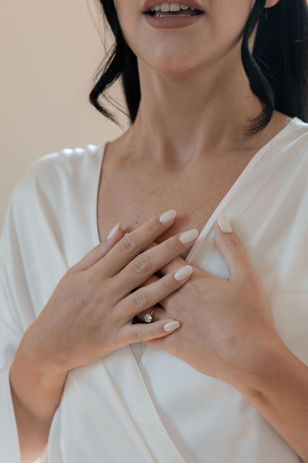 Woman in a white satin robe with manicured nude nails and a diamond ring, resting both hands gently over her chest.