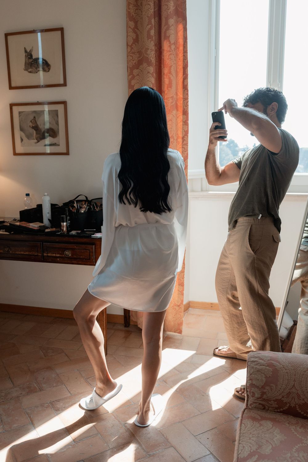 A man in khaki pants photographs a woman in a white robe and slippers in a sunlit room with framed artwork and terracotta tile floors.