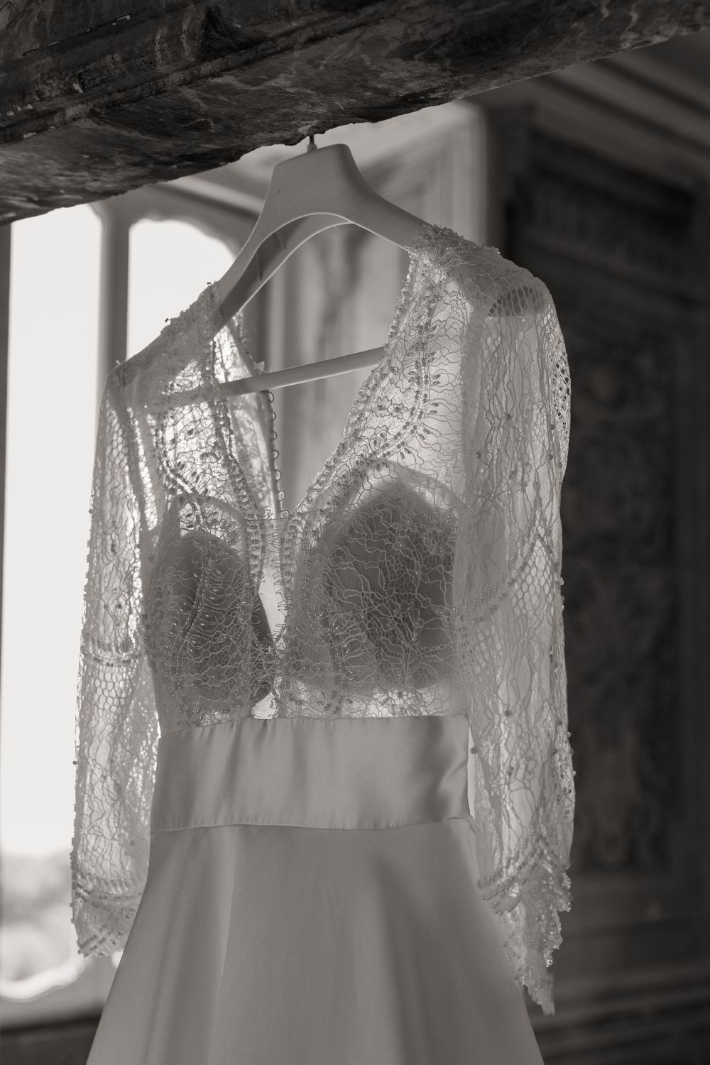 Black and white photo of a lace wedding dress with sheer long sleeves and plunging neckline hanging on a hanger by a window.