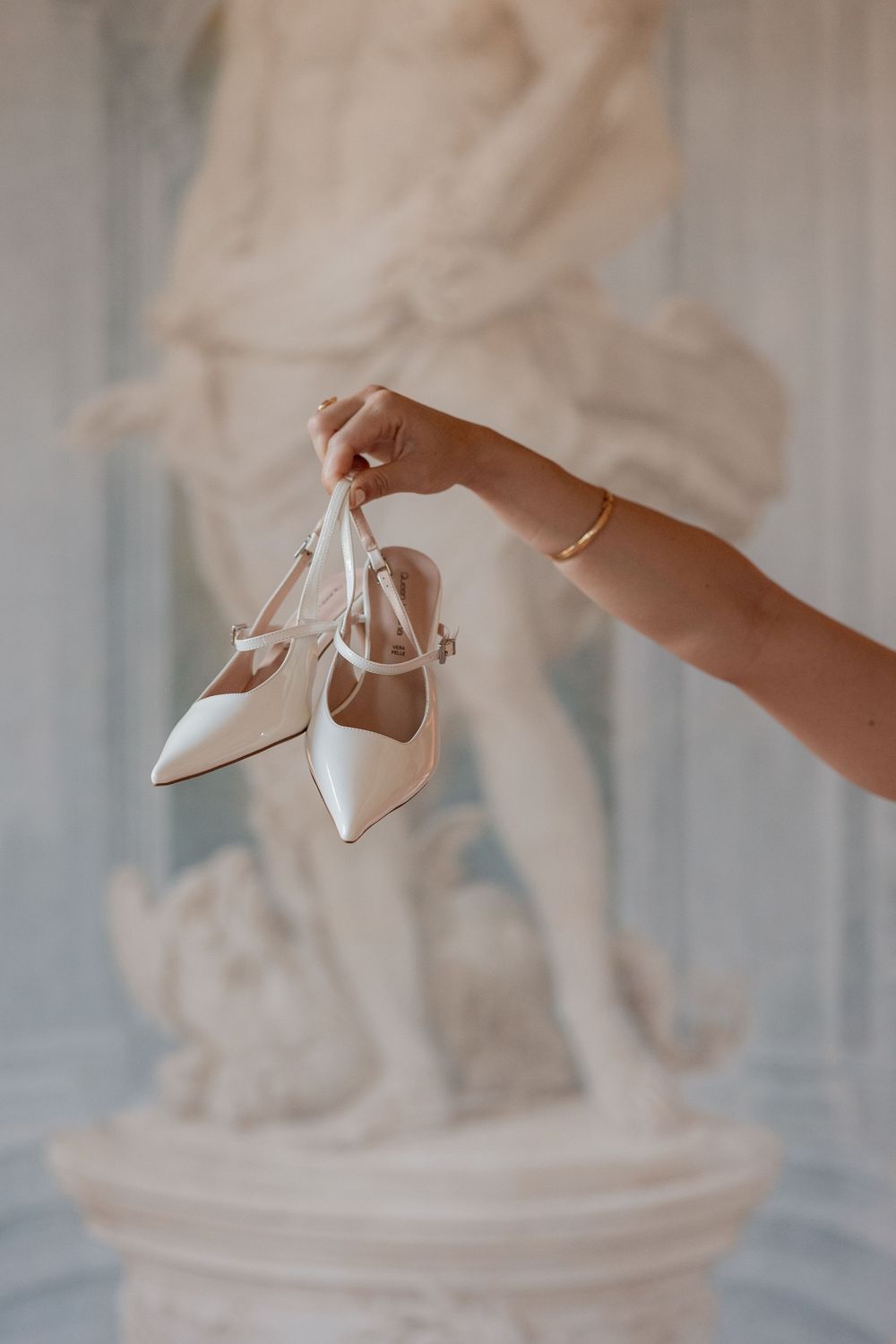 Hand holding a pair of white pointed-toe slingback bridal heels with ankle straps in front of a classical marble statue.