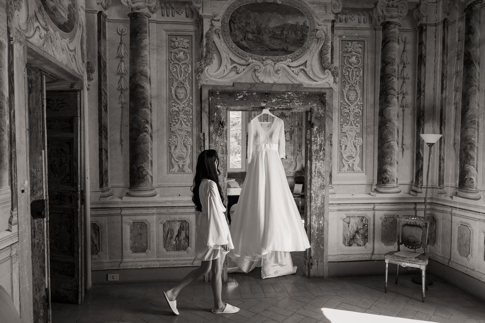 Bride in a white robe walks past a wedding gown hanging in an ornate marble room with frescoed walls in black and white.