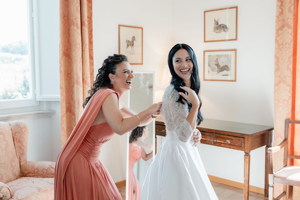 Smiling bride in a lace wedding gown gets help from a bridesmaid in a coral dress while getting ready in a bright room.