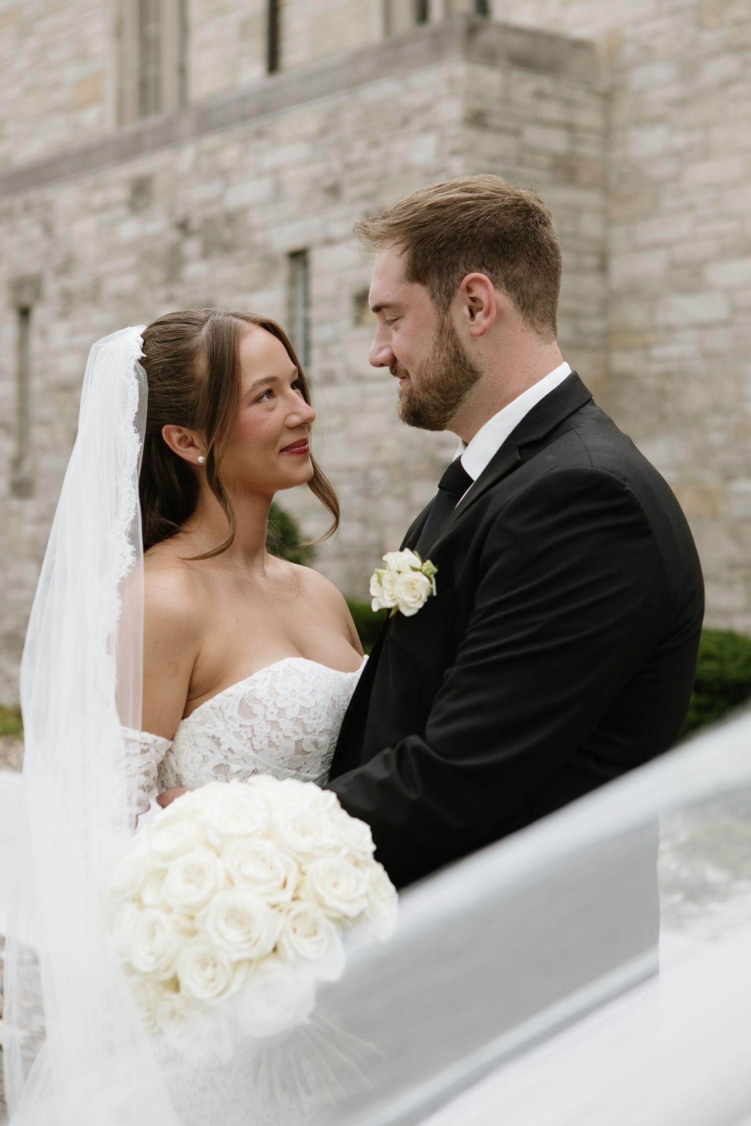 Bride and groom share a romantic moment outdoors, she holds a red and white cascading bouquet, both in elegant wedding attire.