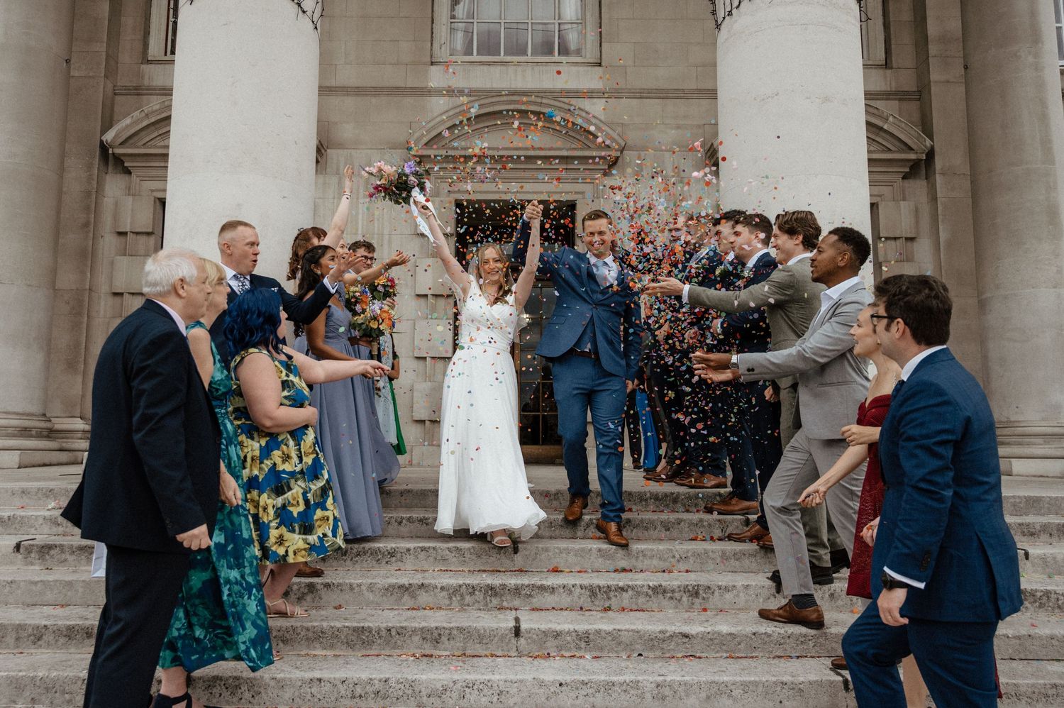Wedding celebration with guests throwing confetti at historic columned building entrance.