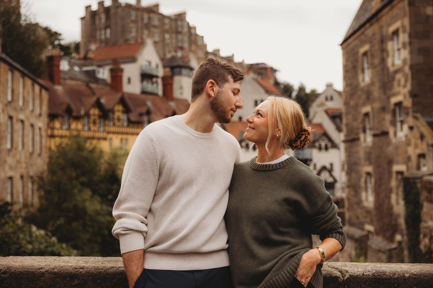 Couple sharing a romantic kiss outdoors against a stone wall with ivy, embracing tenderly in casual attire.