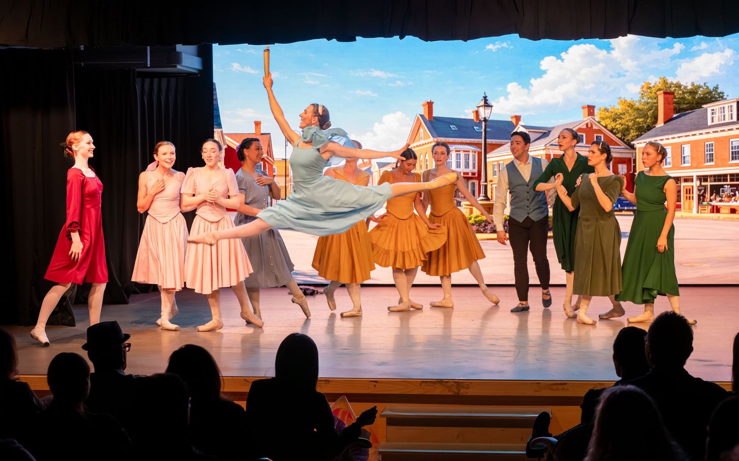 Ballet dancers perform on stage in colorful costumes before a painted small-town backdrop during a live theater production.