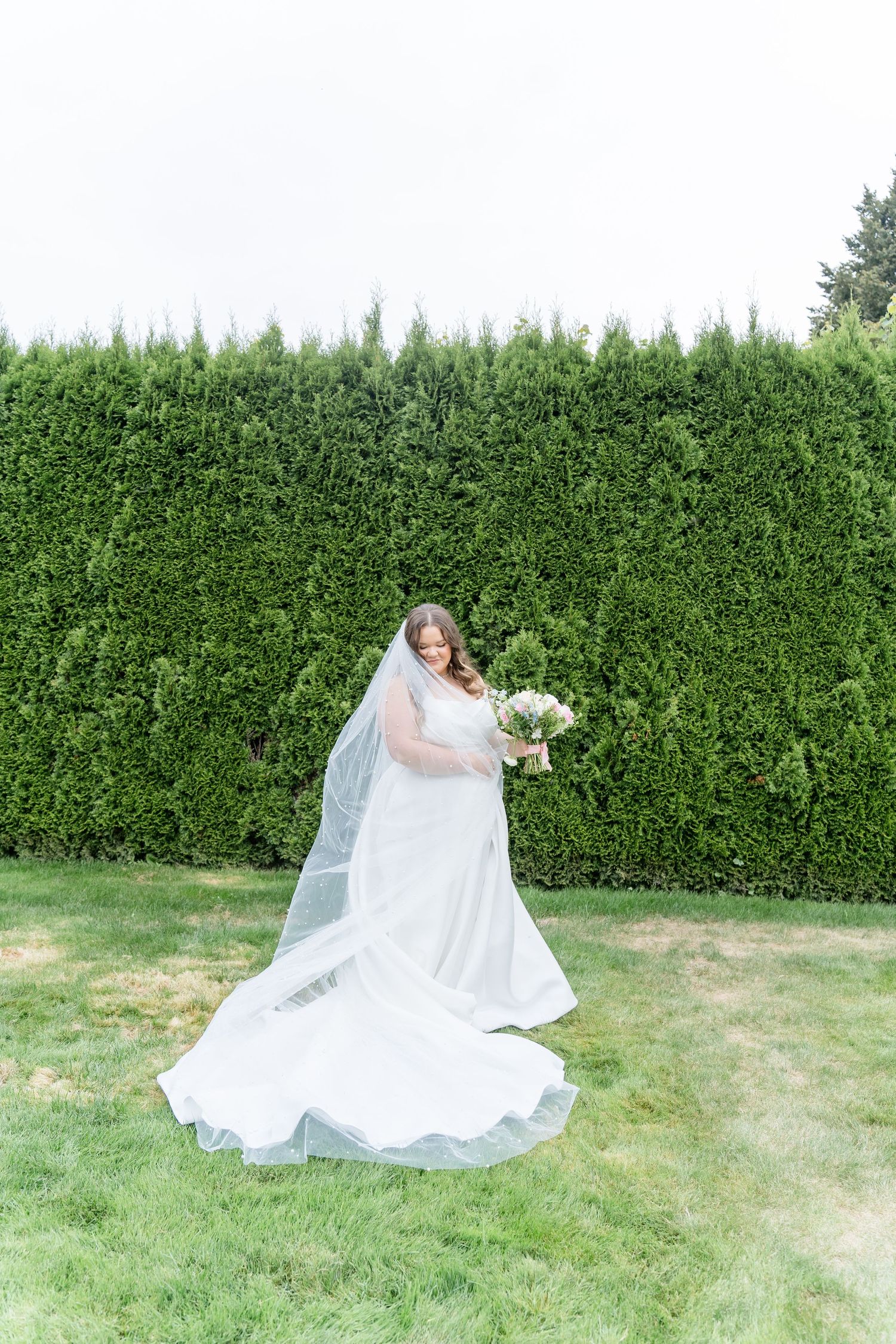 Bride in a flowing white gown and veil holds a bouquet, standing on green grass before a tall hedge backdrop.