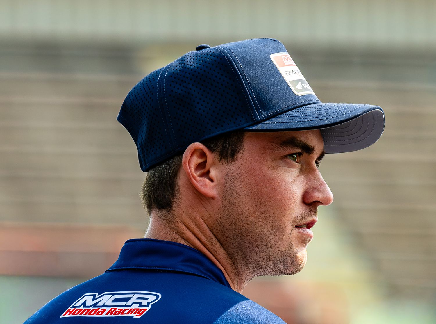 Man in navy MCR Honda Racing cap and shirt stands in profile at a motorsport venue.