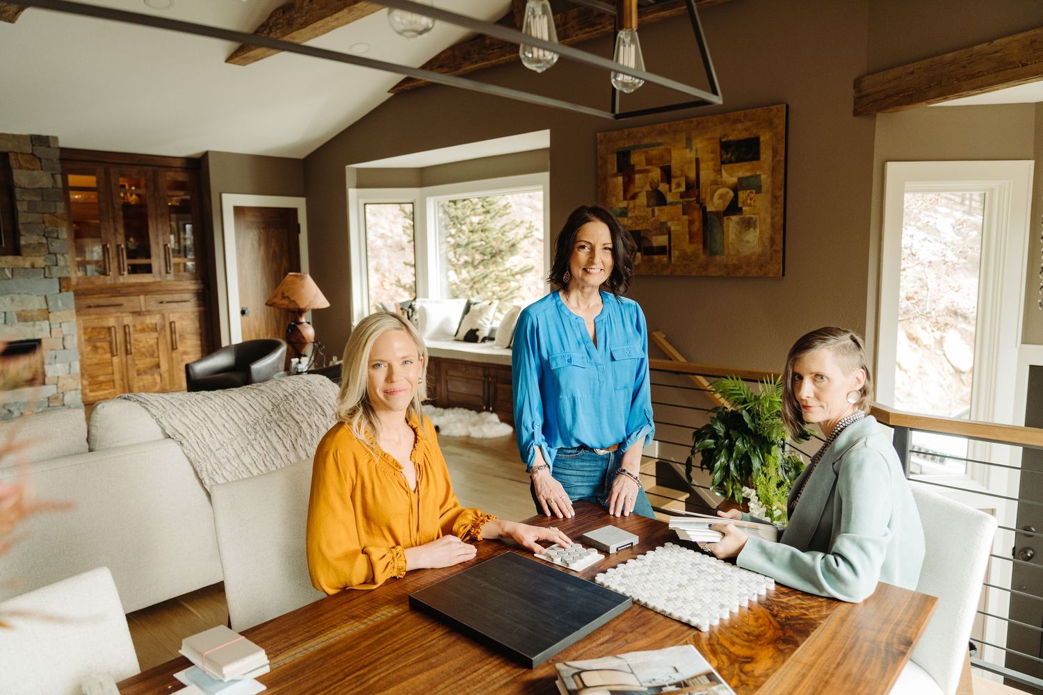 Three women smile around a wooden table with design samples in a cozy home interior setting.