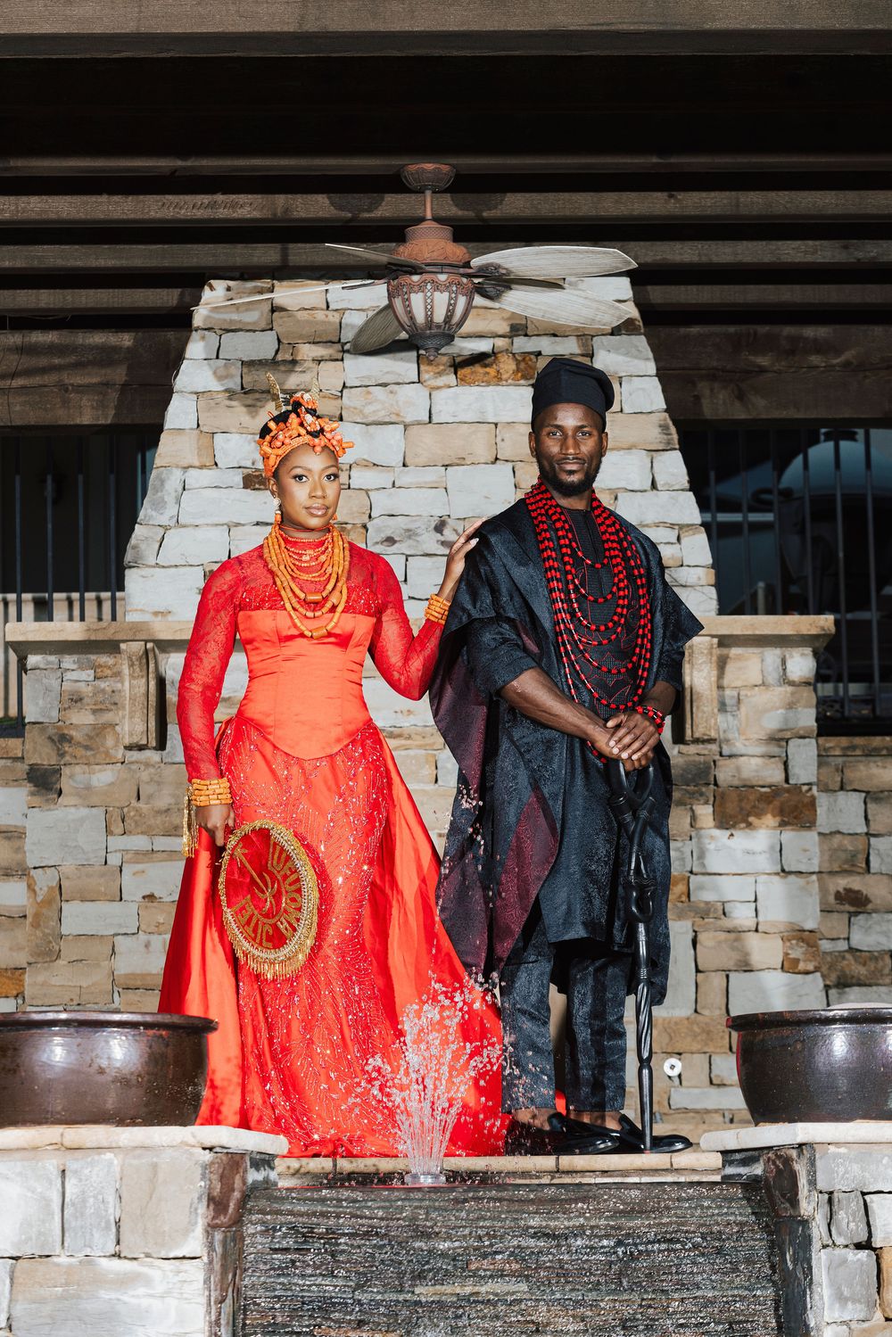 Couple in traditional Nigerian wedding attire, woman in red gown with gold jewelry, man in dark embroidered outfit, stone backdrop.