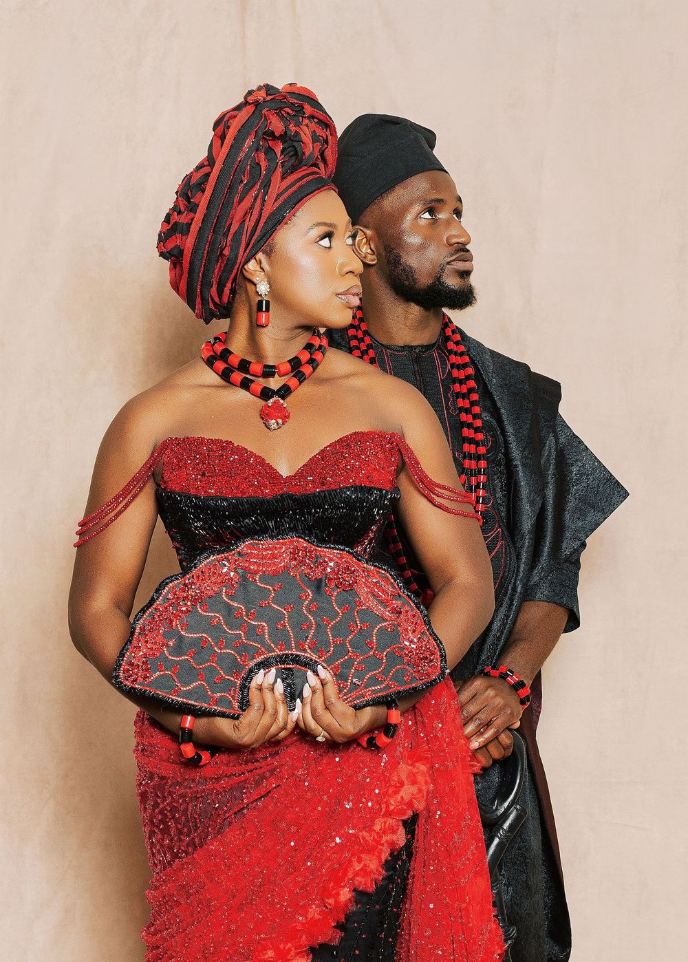 Couple in vibrant red and black traditional African attire, woman holding decorative fan, man in embroidered outfit.
