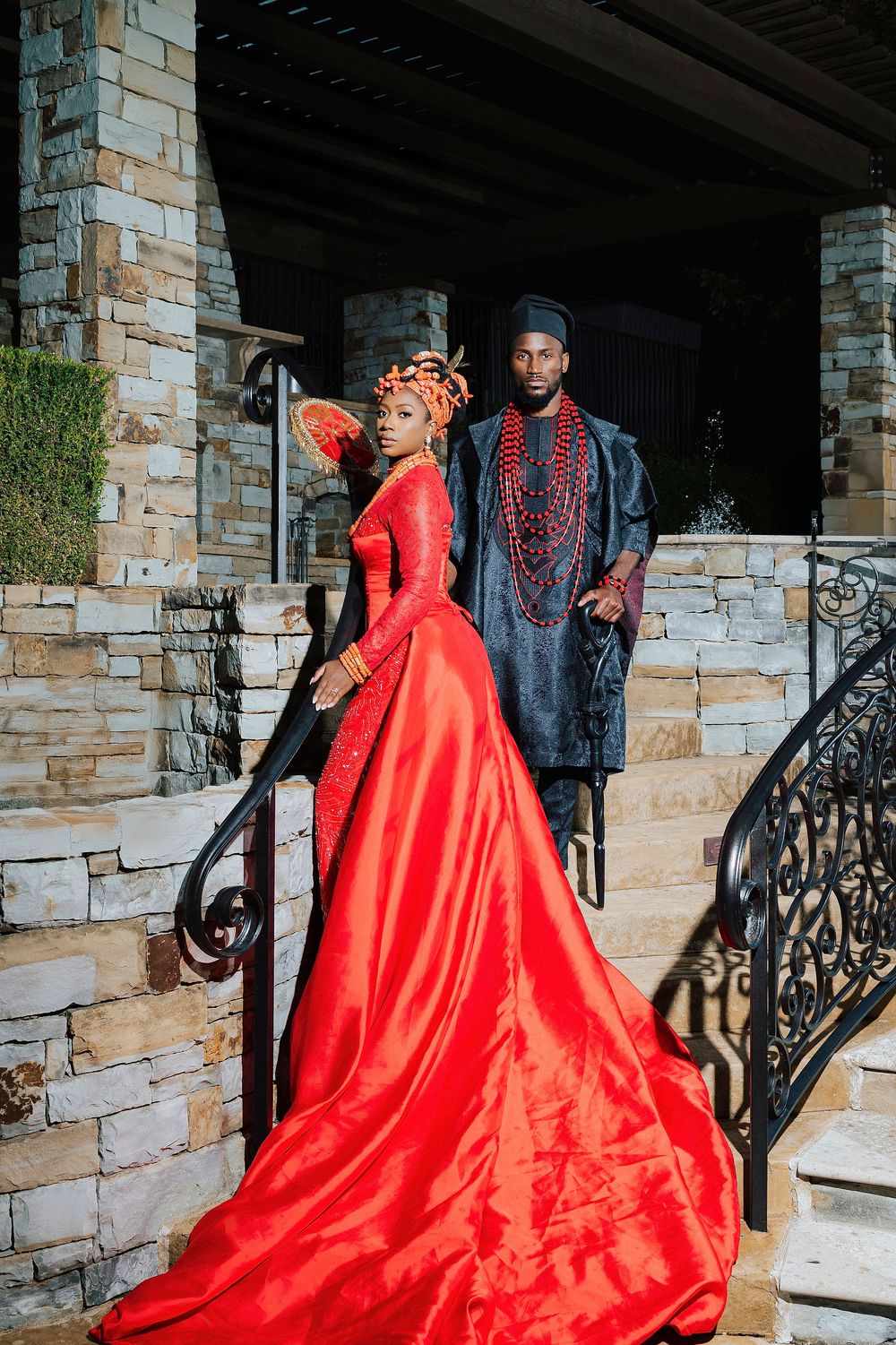 Couple in vibrant African traditional attire, woman in red gown with train, man in navy agbada, posing on stone steps.