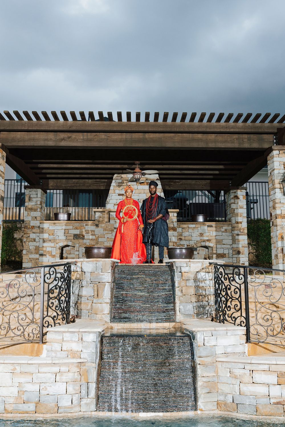 Couple in traditional African attire standing atop a stone waterfall feature under a pergola with dramatic cloudy skies.