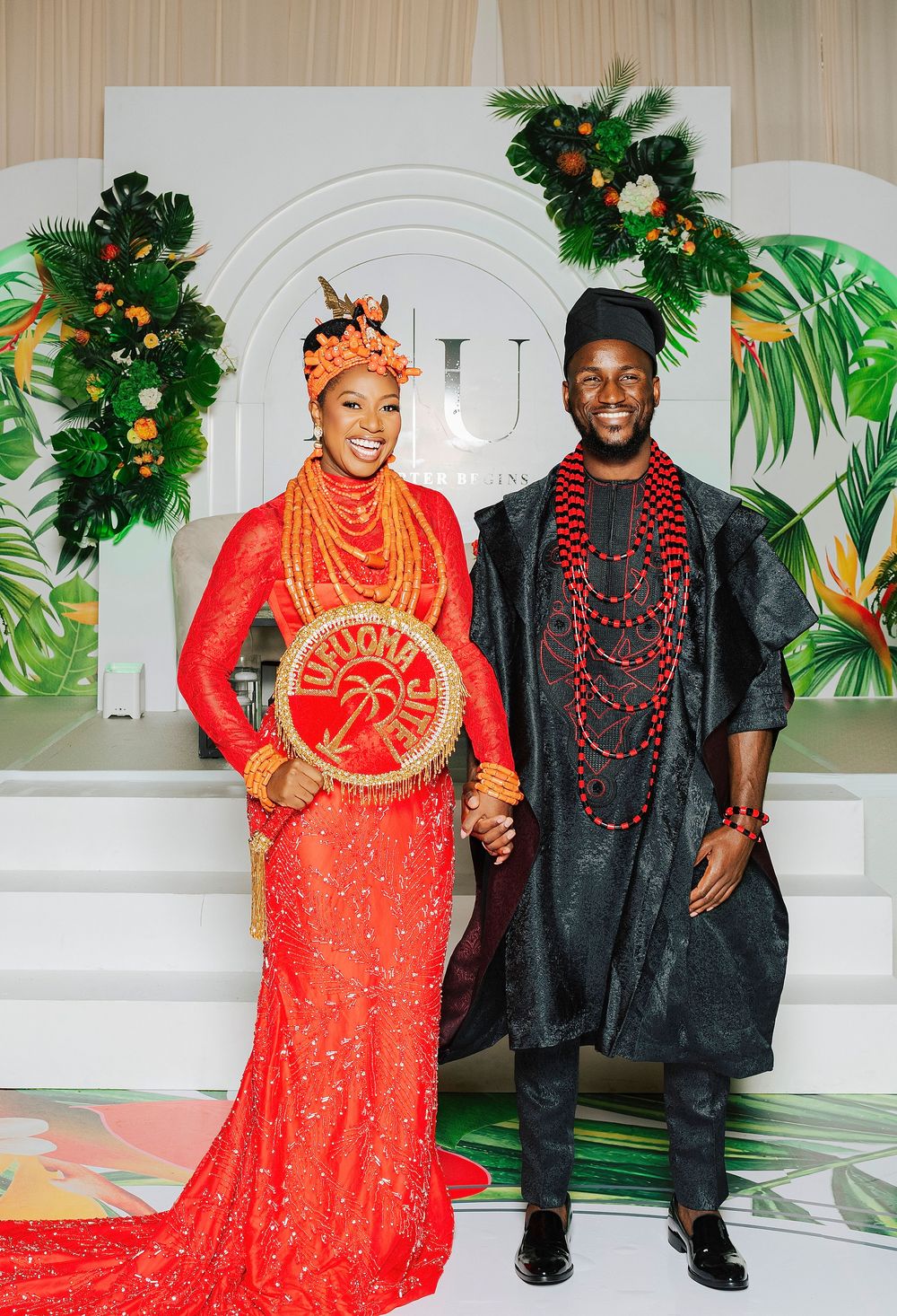 Couple in traditional Nigerian wedding attire, bride in red lace gown with coral beads, groom in black agbada with red beads.