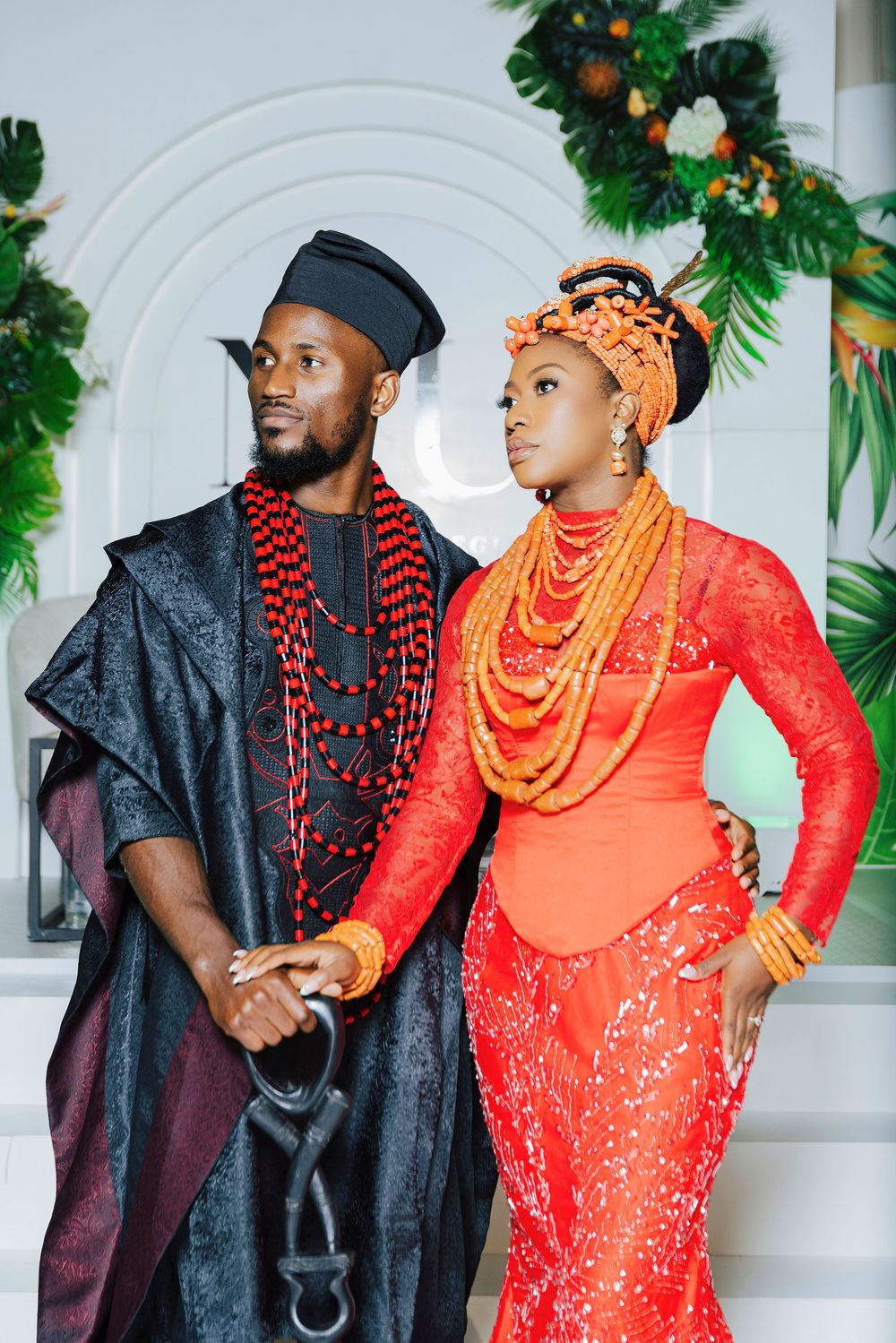 Nigerian couple in traditional Urhobo wedding attire, man in black agbada and woman in orange lace dress with coral beads.