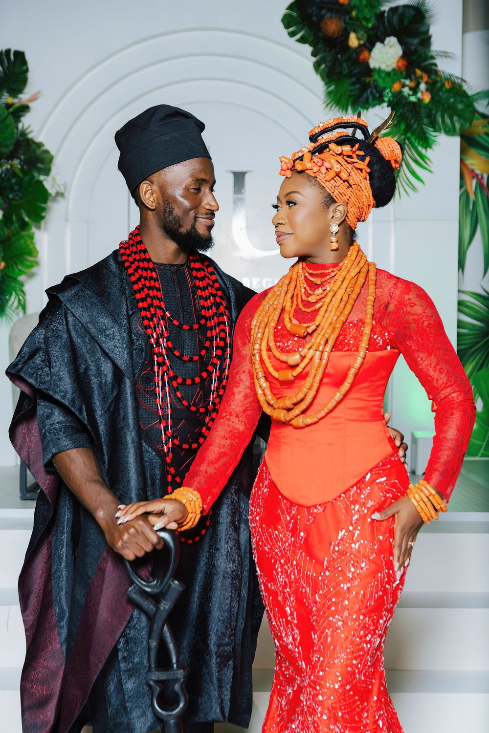 Nigerian couple in traditional Edo attire, man in black agbada, woman in orange lace gown with coral beads.