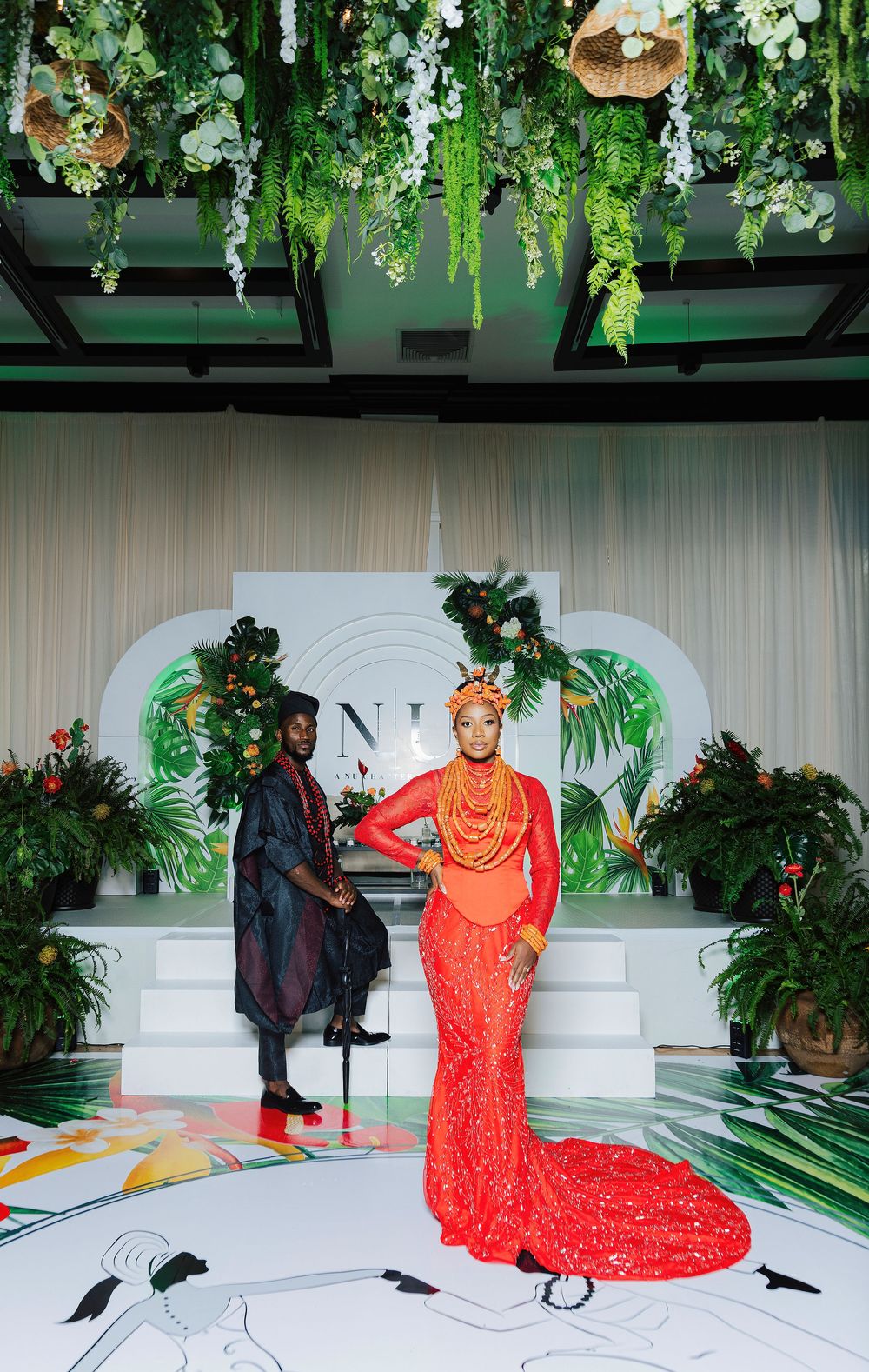 Couple in vibrant traditional African attire pose at an elegant tropical-themed wedding reception with lush greenery decor.