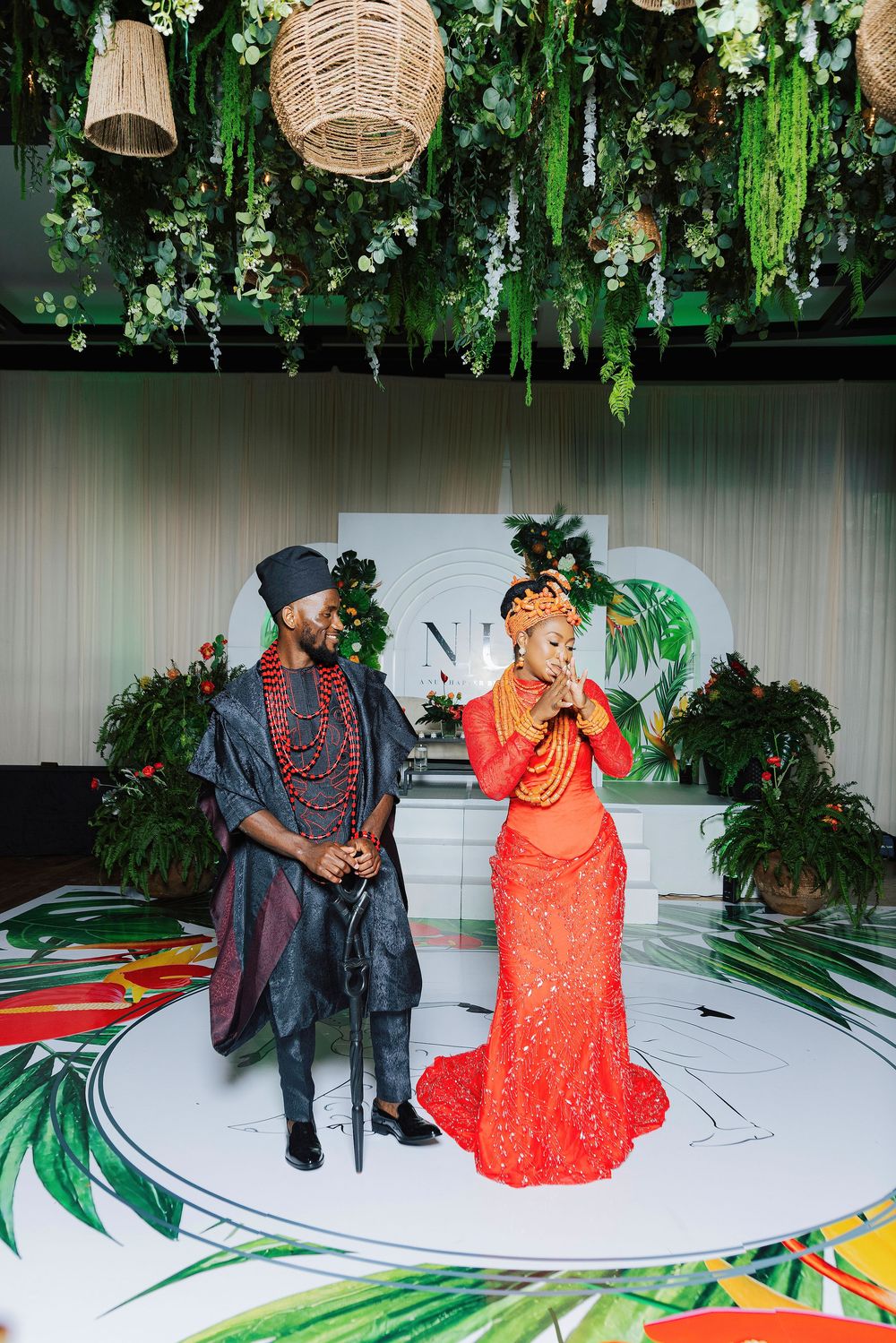 Couple in traditional Nigerian wedding attire stand on decorated stage with tropical floral arrangements overhead.