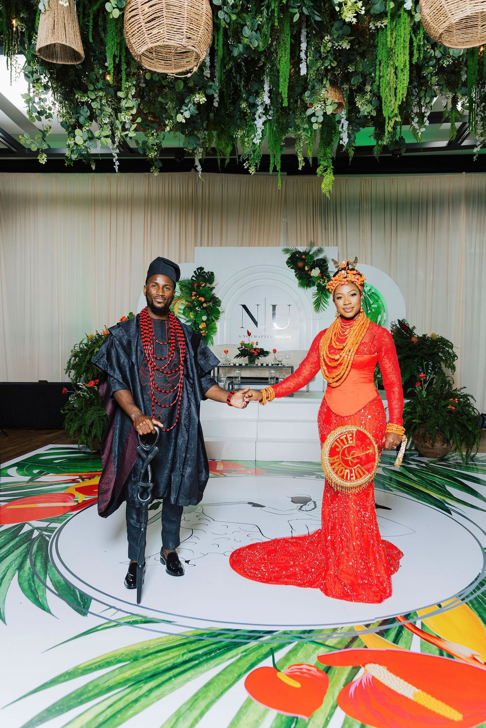 Couple in vibrant Nigerian traditional wedding attire pose on decorated dance floor with tropical greenery overhead.