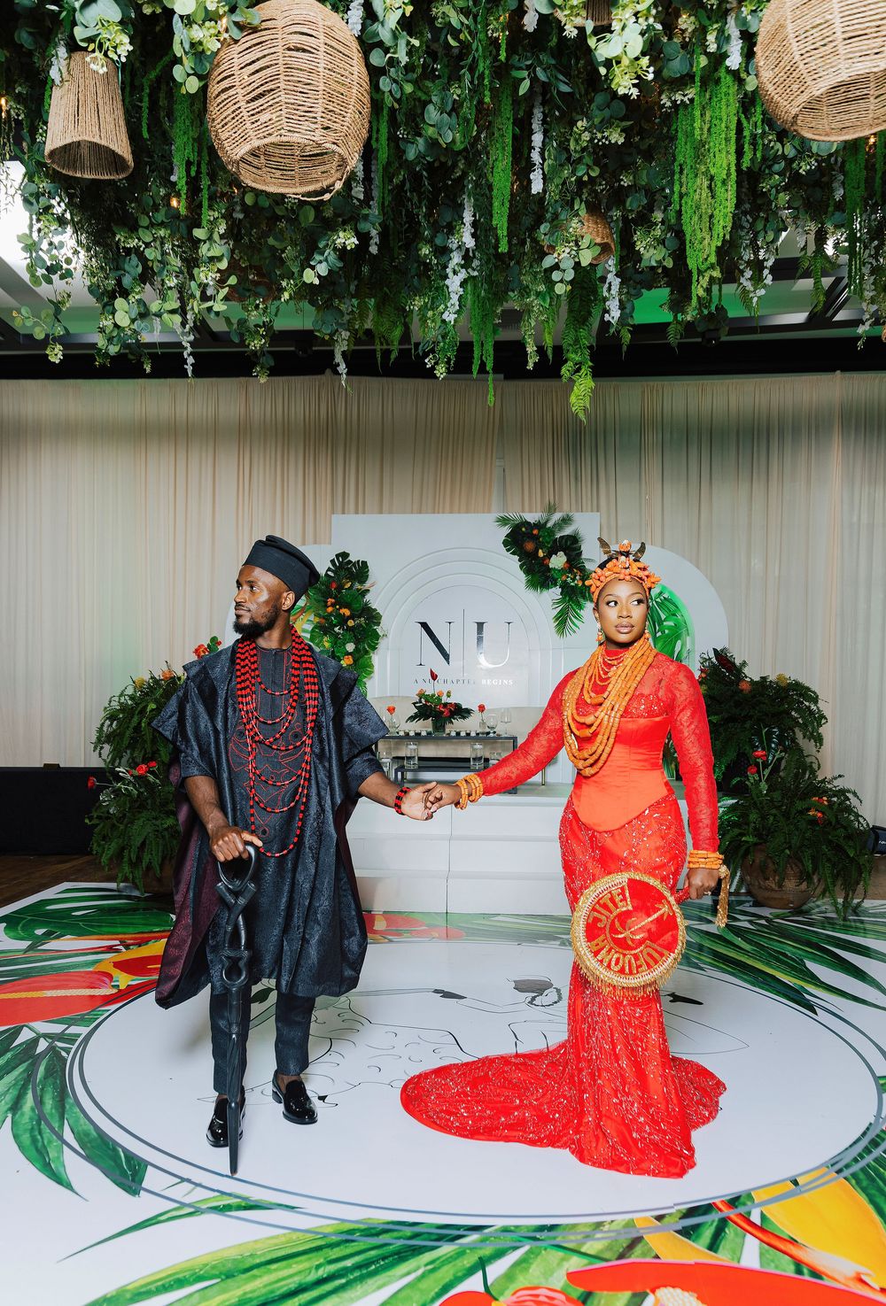 Couple in vibrant red and navy traditional Nigerian wedding attire holding hands at a lush tropical-themed ceremony.