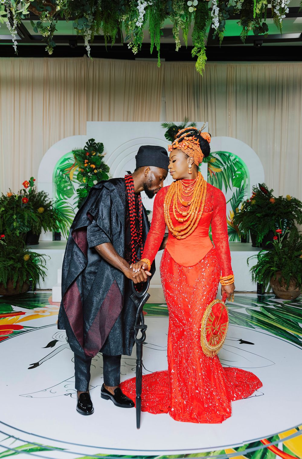Nigerian couple in traditional wedding attire, bride in red lace gown with coral beads, groom in black agbada, sharing romantic moment.