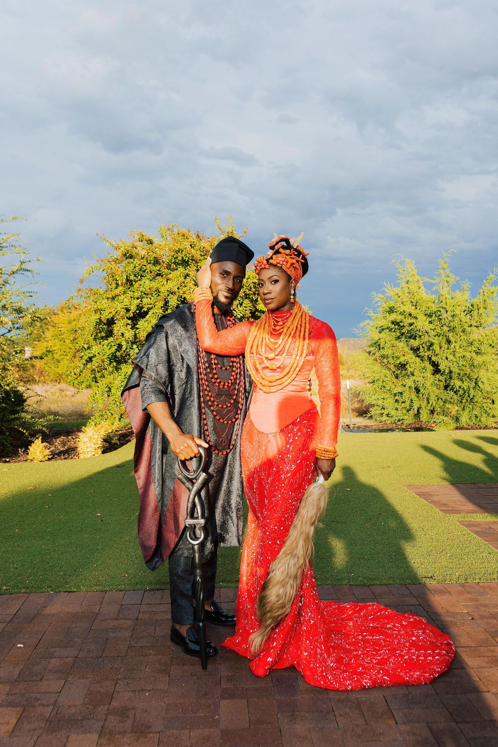 Couple in vibrant traditional African attire posing outdoors, woman in red sequin gown with orange headwrap, man in gray embroidered outfit.