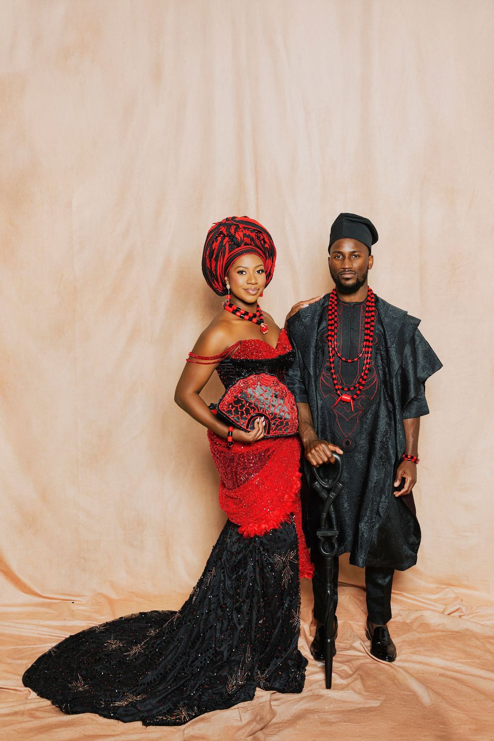 Couple in traditional Nigerian attire, woman in red lace gown with gele headwrap, man in black agbada outfit.