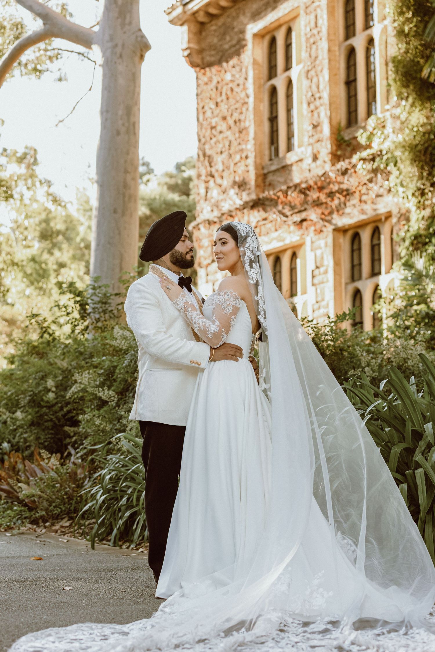 Sikh groom in white suit and turban stands with bride in lace wedding gown and veil outside historic stone mansion.