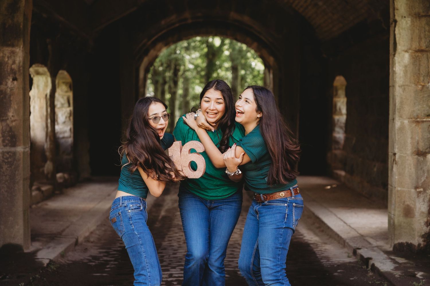 Three young women laughing and posing together under a stone arch tunnel, one holding a number 16 sign.