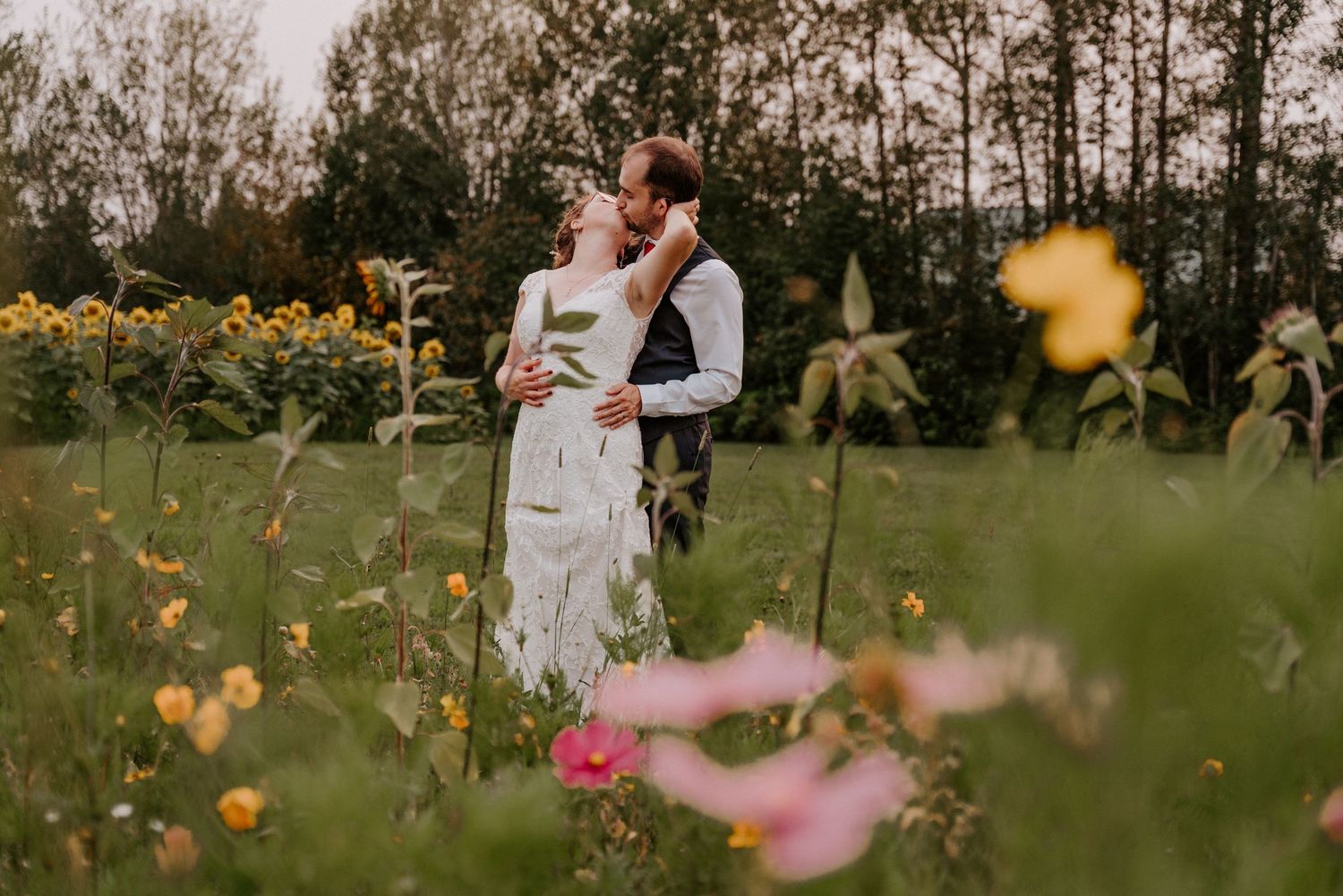 A couple shares a romantic kiss surrounded by wildflowers and sunflowers in a lush green field on their wedding day.