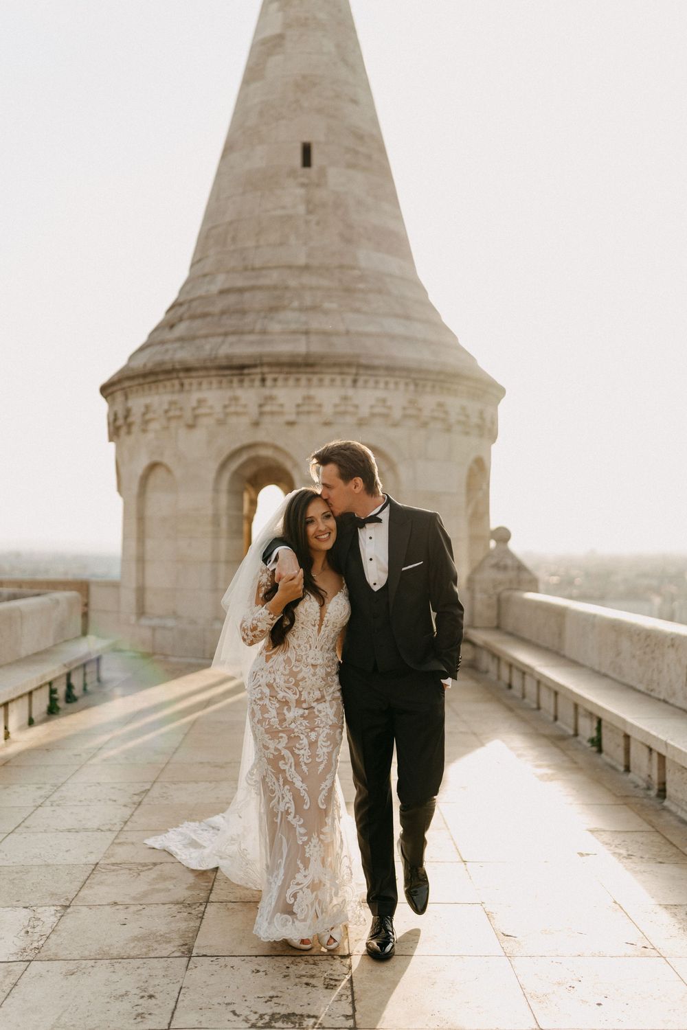 Groom kissing bride near stone tower at Fishermans Bastion during golden hour.