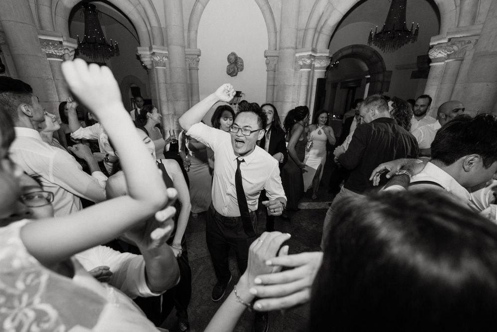 Groom dancing energetically with hands raised among wedding guests in black and white reception photo.