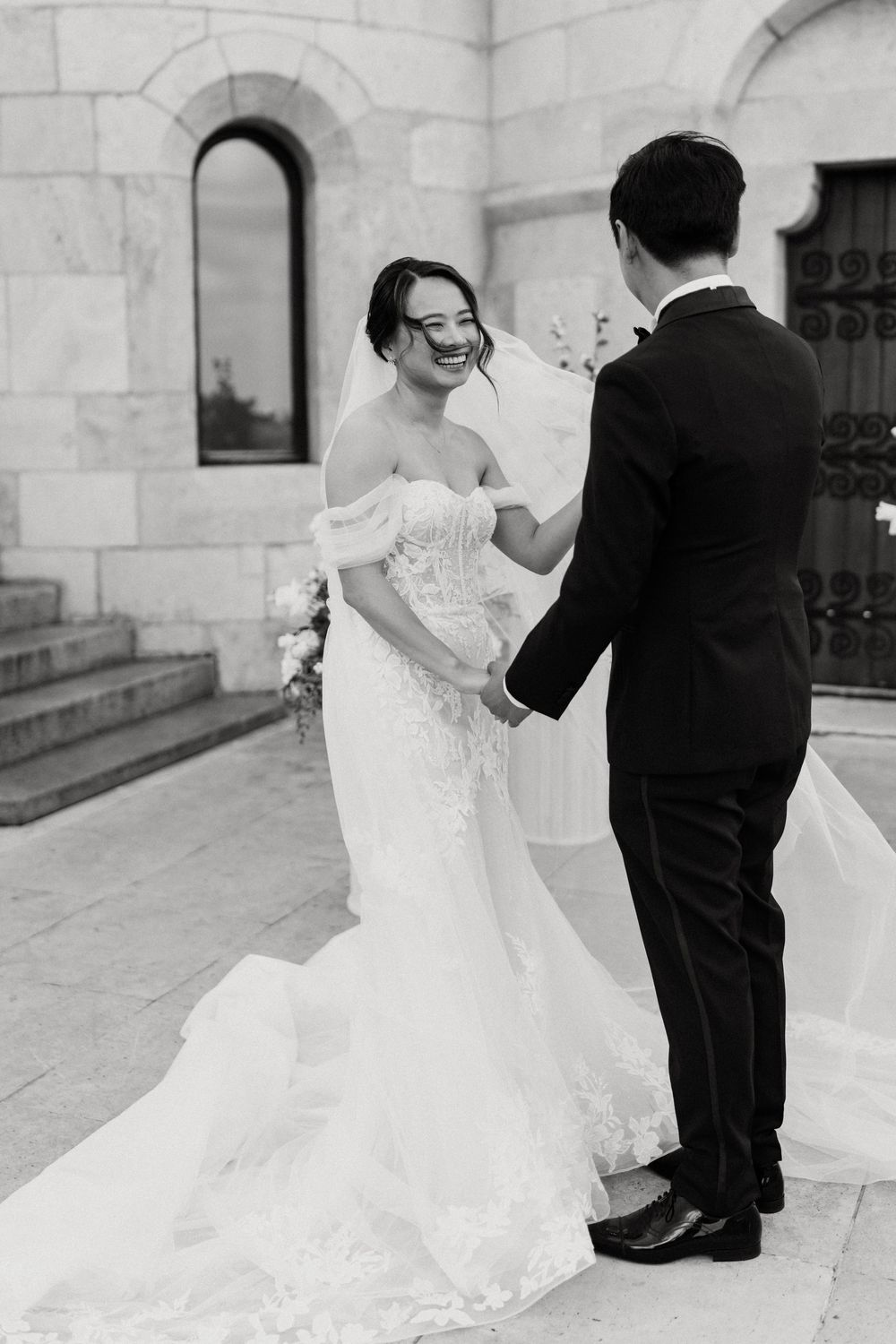 Bride in off-shoulder lace gown laughing while holding groom's hands outside stone church in black and white.