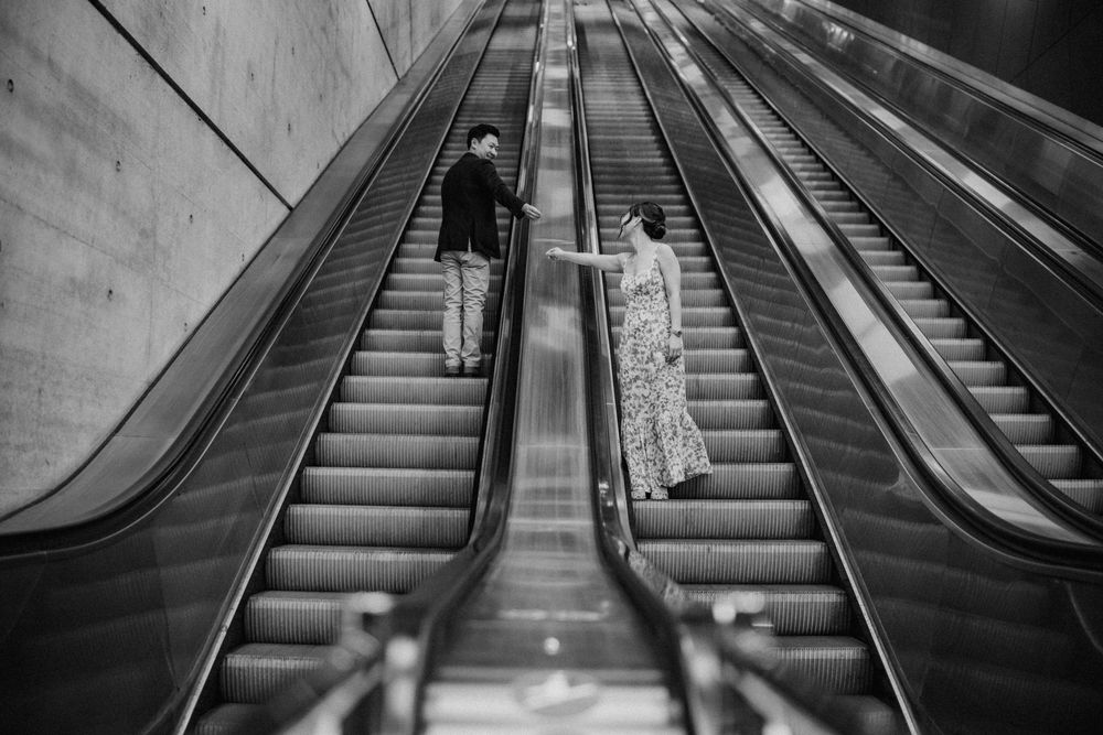 Black and white photo of couple holding hands on opposite escalators.