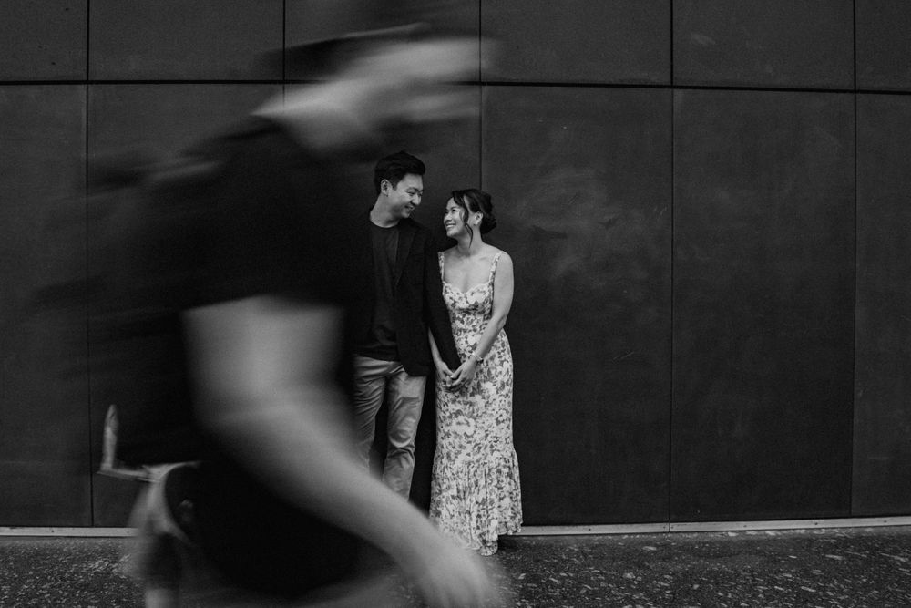 Black and white street photo of couple smiling with blurred pedestrian passing.