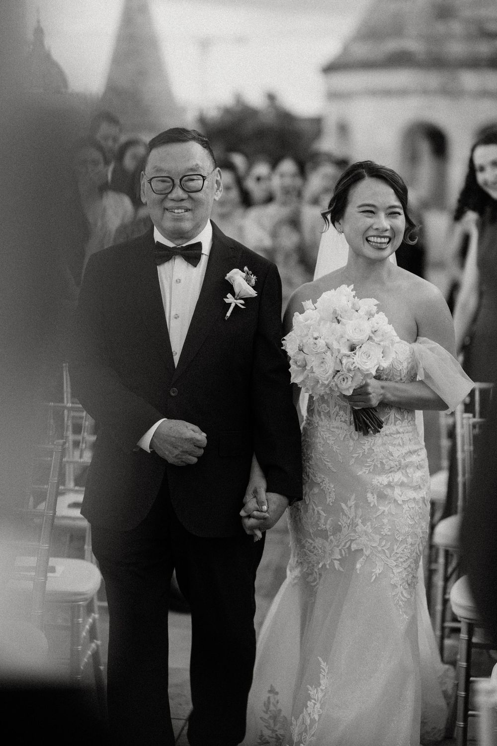 Smiling bride in lace gown walking down aisle with father in tuxedo holding white bouquet at outdoor ceremony.