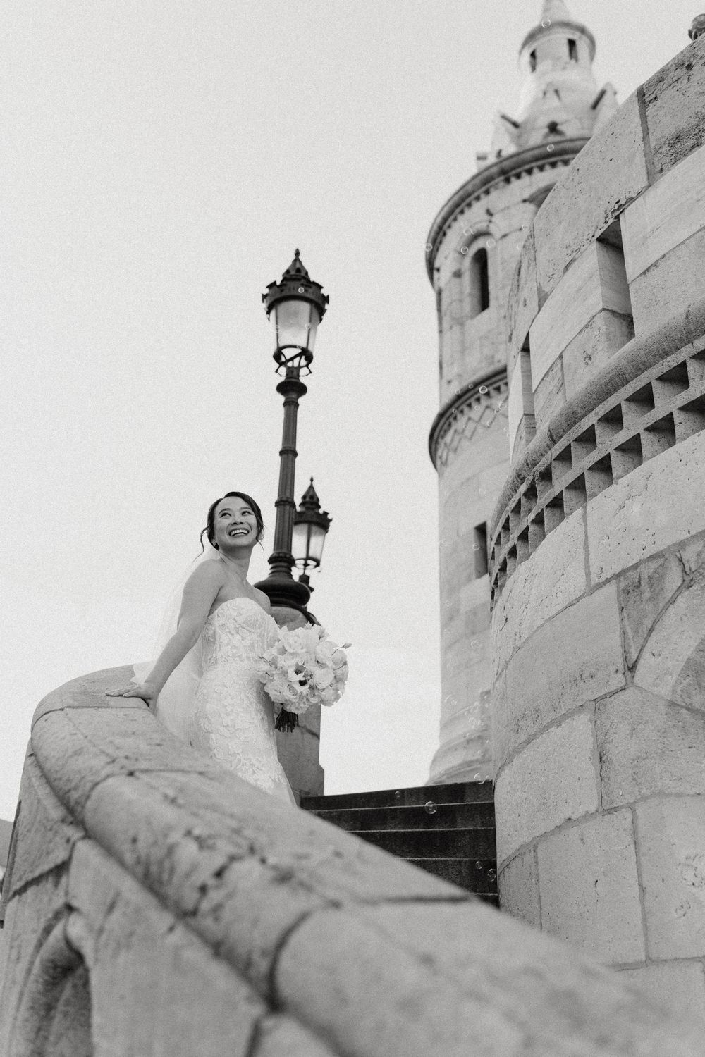 Smiling bride in lace gown holds bouquet on stone steps beside a lamp post and historic castle tower.