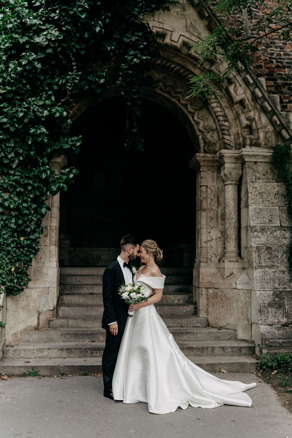 Bride and groom share a romantic kiss before an ivy-covered stone archway with grand steps behind them.