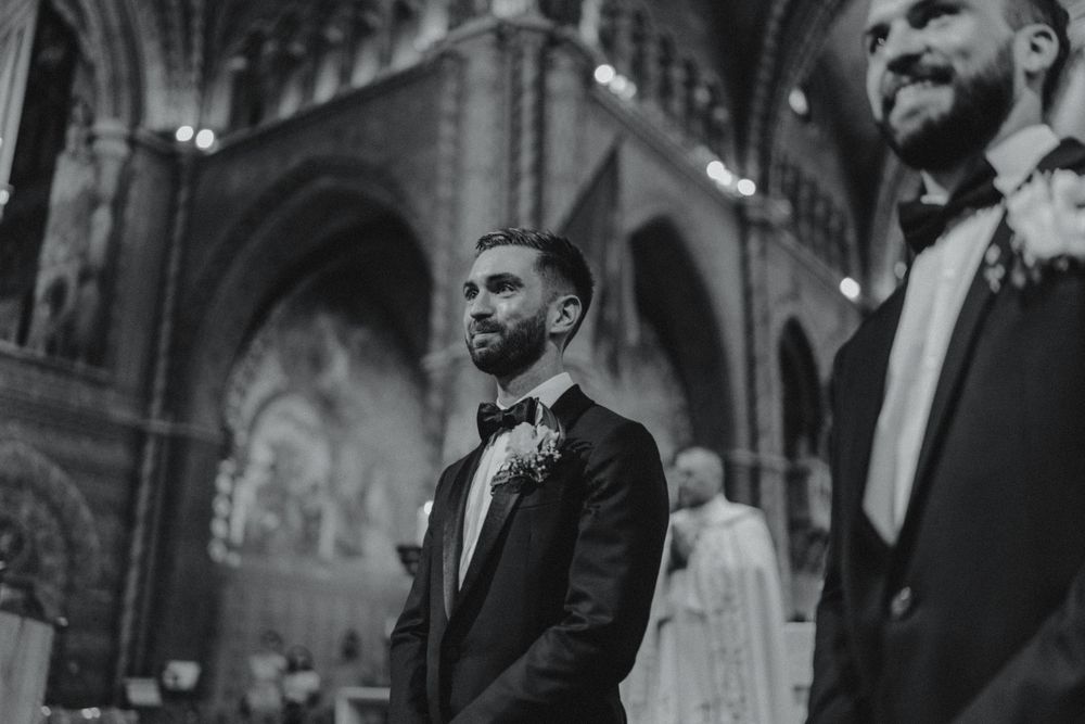 Emotional groom in tuxedo smiles at the altar inside a stunning Gothic cathedral in black and white.