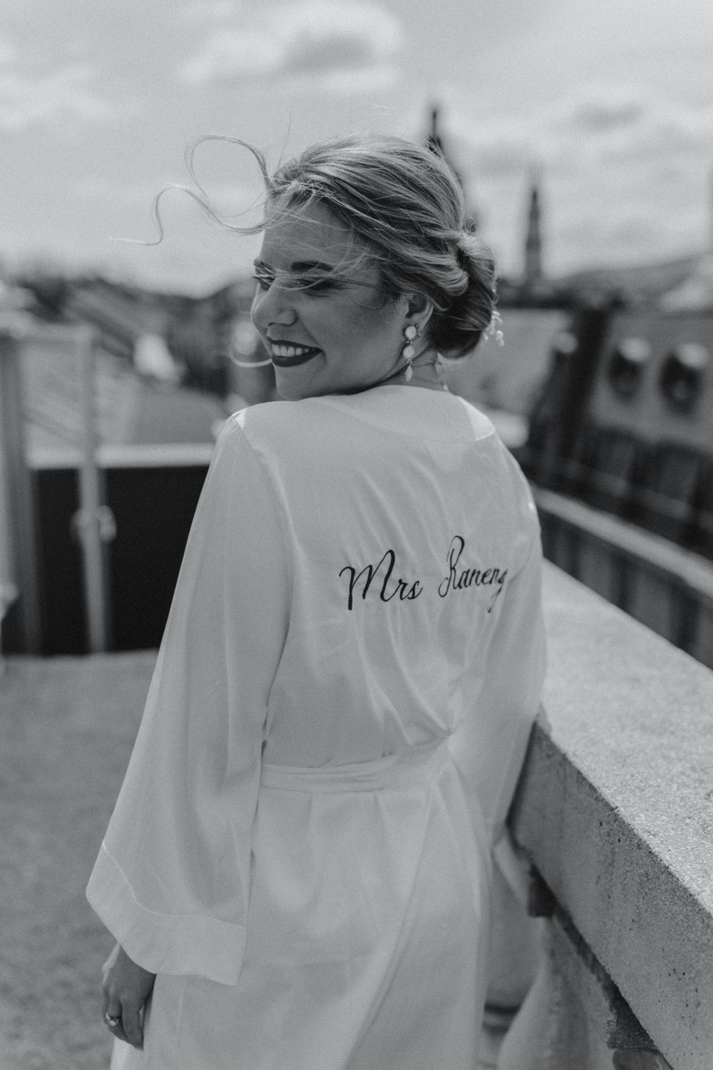 Bride in personalized Mrs. surname white robe laughing on rooftop with city skyline in background.