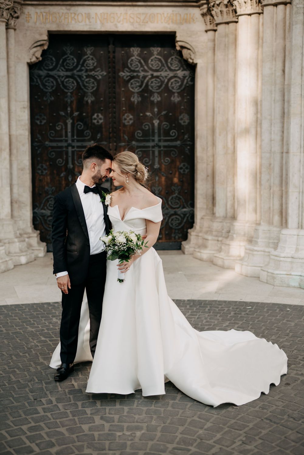 Wedding couple sharing romantic forehead touch before ornate iron doors of historic stone cathedral.