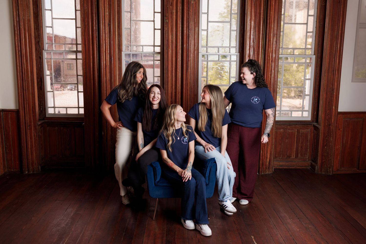 Five young women in navy blue shirts pose together laughing near large wooden windows in an elegant room.
