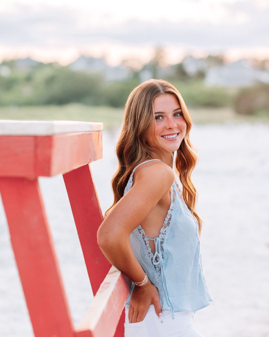 A smiling young woman with auburn hair leans against a red and white striped post near a beach at sunset.