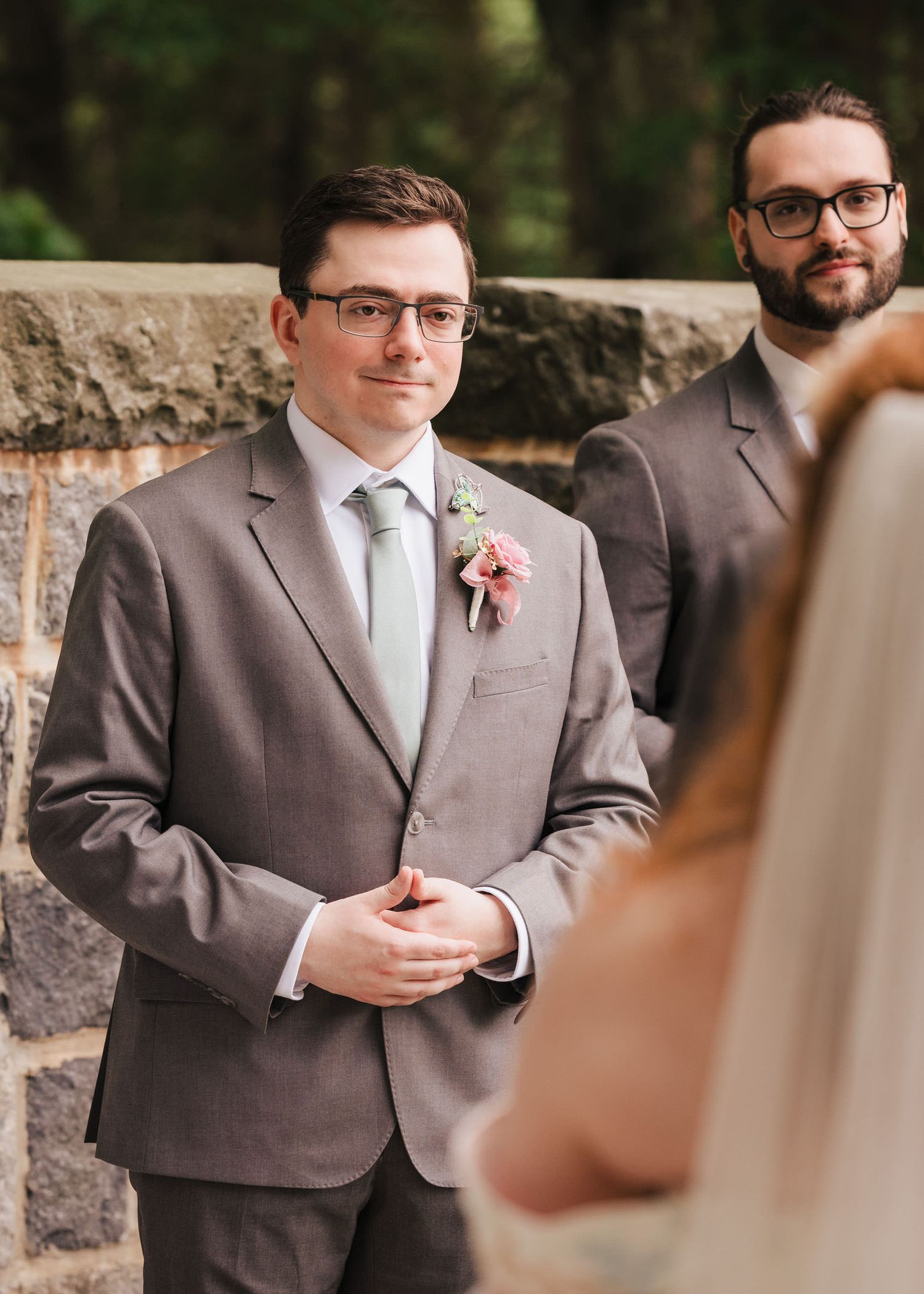 Best man in a gray suit and sage tie watching the ceremony, second best man visible in the background.