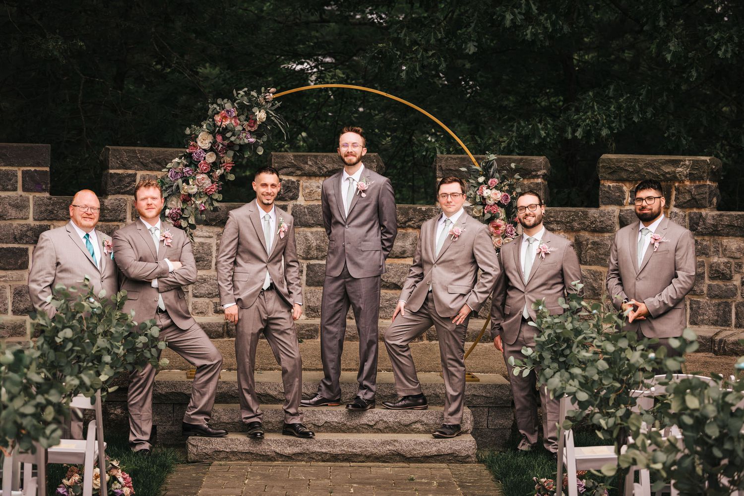 Groom and groomsmen posing together at the ceremony arch at courtyard in Searles Castle, in front of a stone wall and floral arbor.